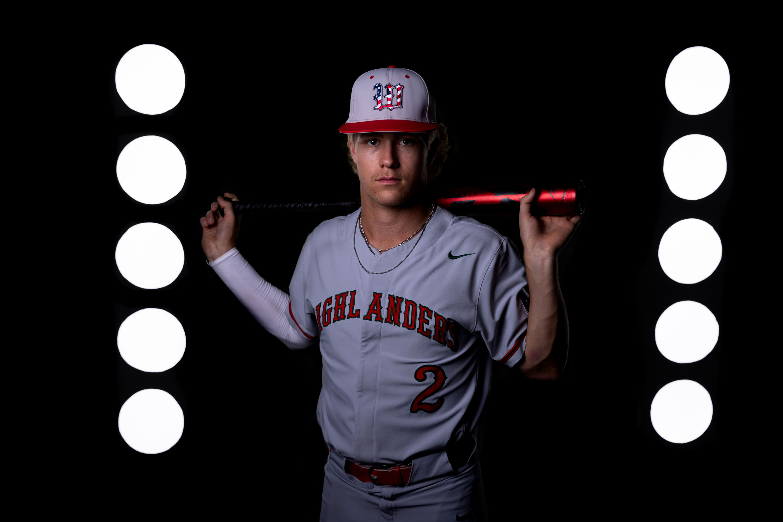 The Woodlands High School Highlanders baseball player standing with bat on shoulders and stadium lights behind him.