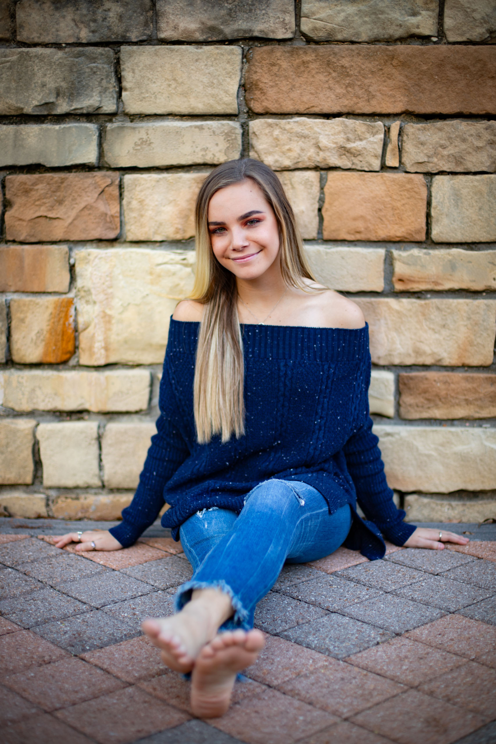Girl in blue sweater and jeans sitting against brick wall.